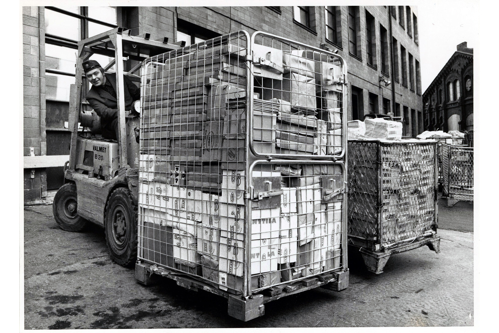 Postin 1970-luvun työasussa oleva mies siirtää paketteja täynnä olevaa tavarakärryä haarukkatrukilla.