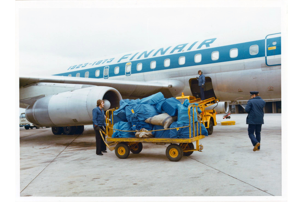 Lentopostisäkeistä täysinäinen kärry lastausaukon luona Finnairin koneen edessä kolmen työmiehen työskennellessä lähettyvillä.