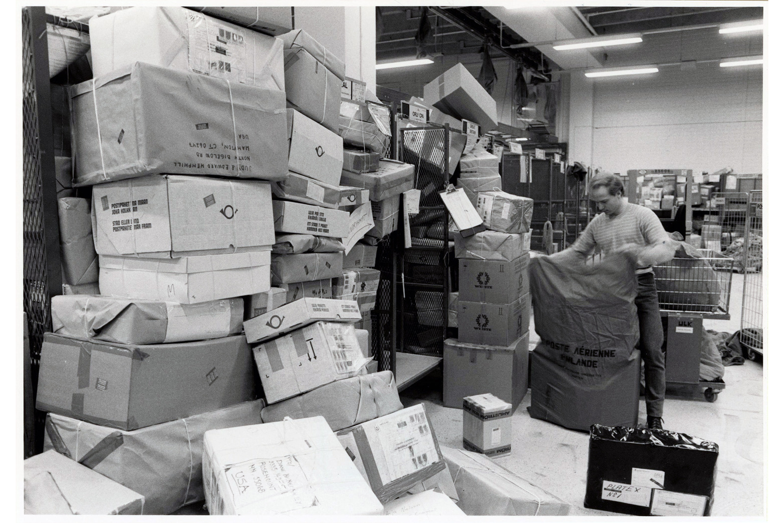 Kasoittain isoja postipaketteja hallin seinustoilla työntekijän avatessa postisäkkiä.