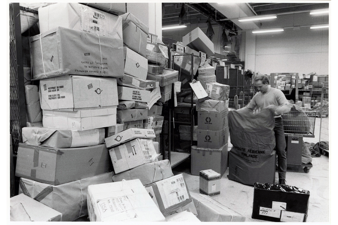 Kasoittain isoja postipaketteja hallin seinustoilla työntekijän avatessa postisäkkiä.