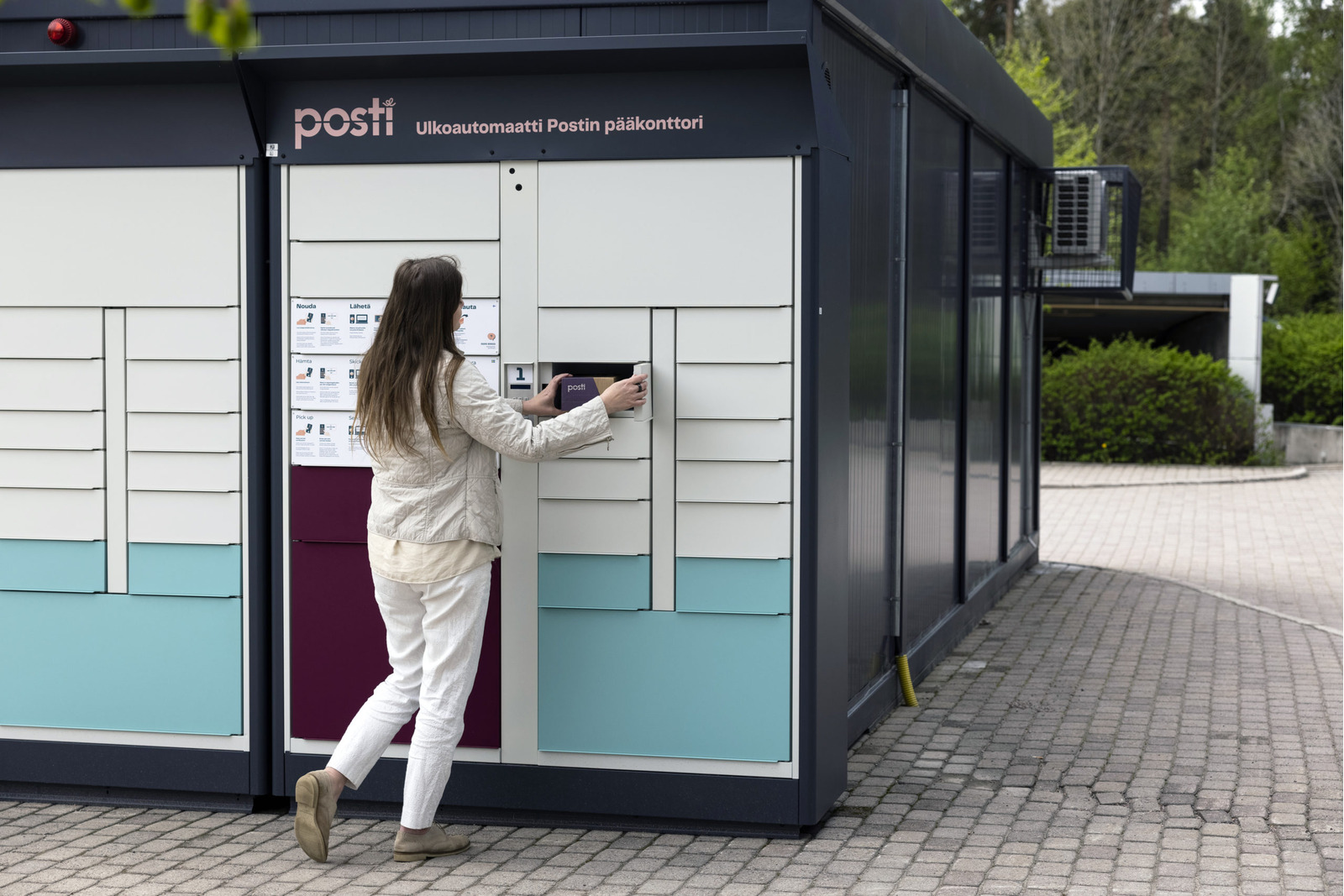 Postin ulkoautomaatilla nainen ottamassa pakettia lokerikosta kesäaikaan.