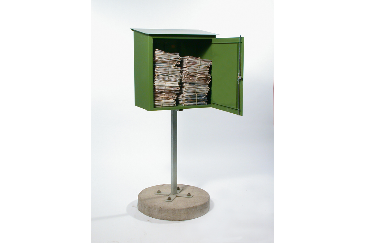 yhdellä jalalla betonirenkaan päällä seisova vihreä metallinen lukittava laatikko auki, jossa sisällä yhteen narulla sidottuja sanomalehtinippuja.