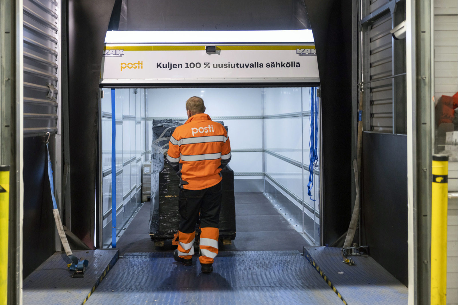 Postin työasussa oleva mies siirtää täysinäistä tavarakärryä Postin sähkökuorma-autoon.