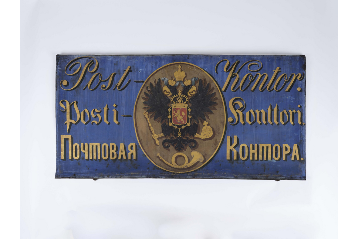 Vanha sinipohjainen postin kyltti, jossa lukee postikonttori kolmella eri kielellä kellertävällä, keskellä kaksipäinen kotka.