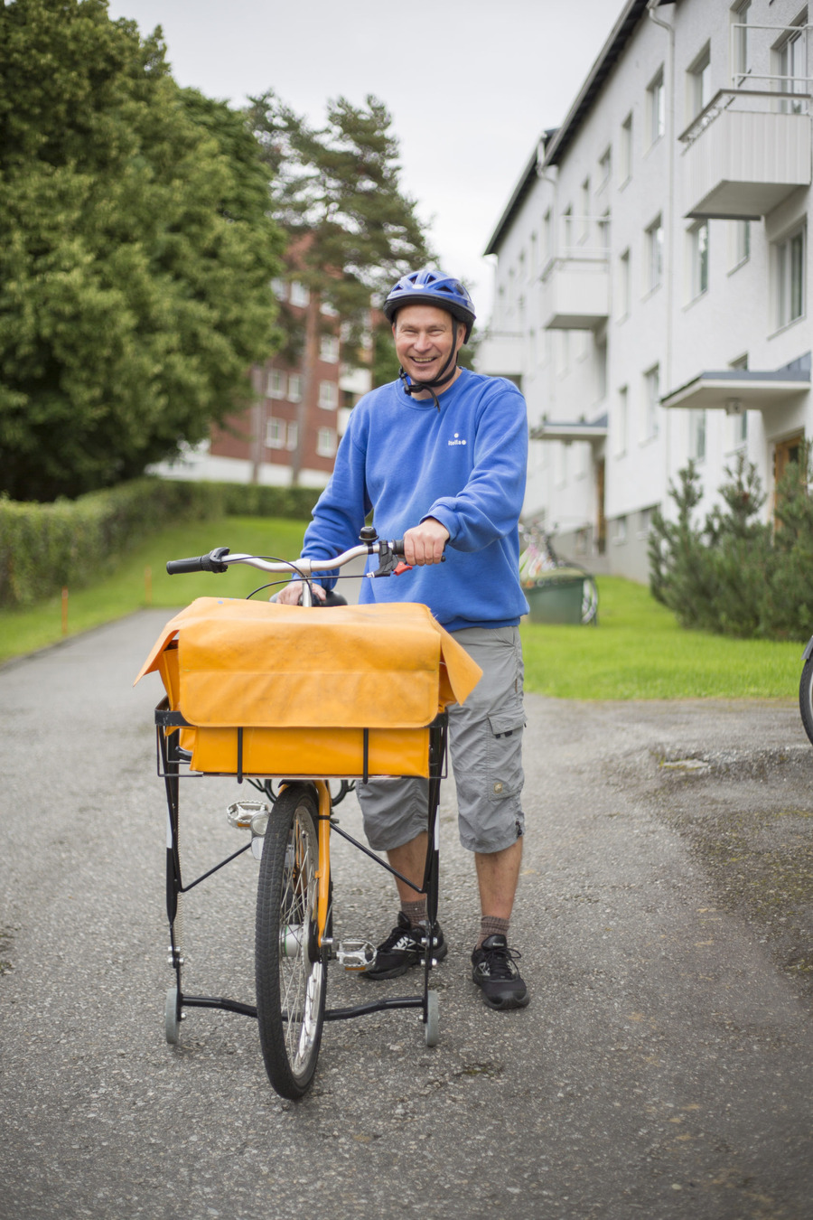 Postinjakaja jakelupyöränsä kanssa 2007. Hänellä on päällään sininen kypärä ja pusero sekä polvipituiset harmaat housut.