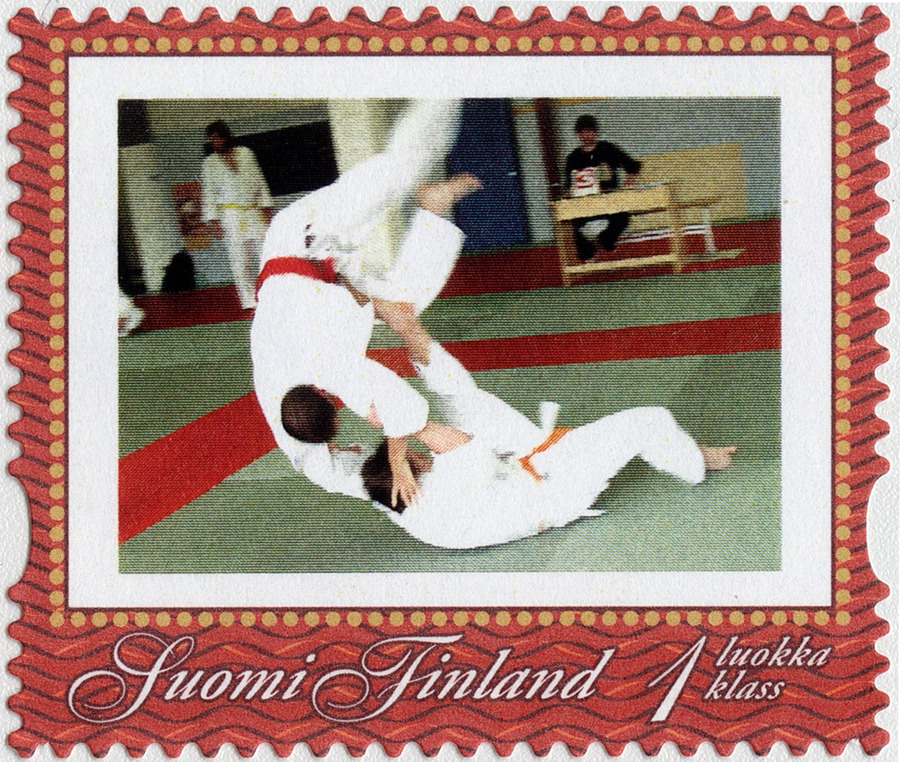 Omakuvapostimerkin kuvassa toinen judoka on maassa ja toinen ilmassa. Maassa olevalla oranssi vyö, ilmassa olevalla punainen.