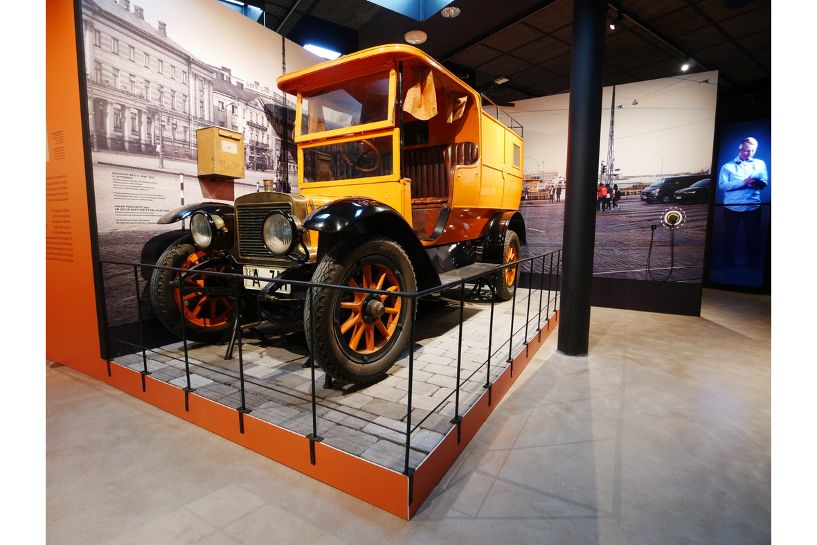 Näyttelytila, jossa on kello kahdessatoista vinossa nupukivien päällä aidattuna vanha postiauto. Se sijaitsee kahden seinäpinnan nurkasssa. Sen vieressä seinällä vasemmalla on vanha kirjelaatikko. Suoraan edessä on pylväs. Pylvään oikealla puolella seinällä, sen reunassa on kuuloke kehien keskellä. Seinän päädyn takaa pilkistää kauempana oleva näyttö, jossa on mies kameran kanssa. Seinillä on kuvasuurennus Helsingin kauppatorista niin, että vasemmanpuoleisessa on mustavalkoinen ja oikeallapuolella värikuva.