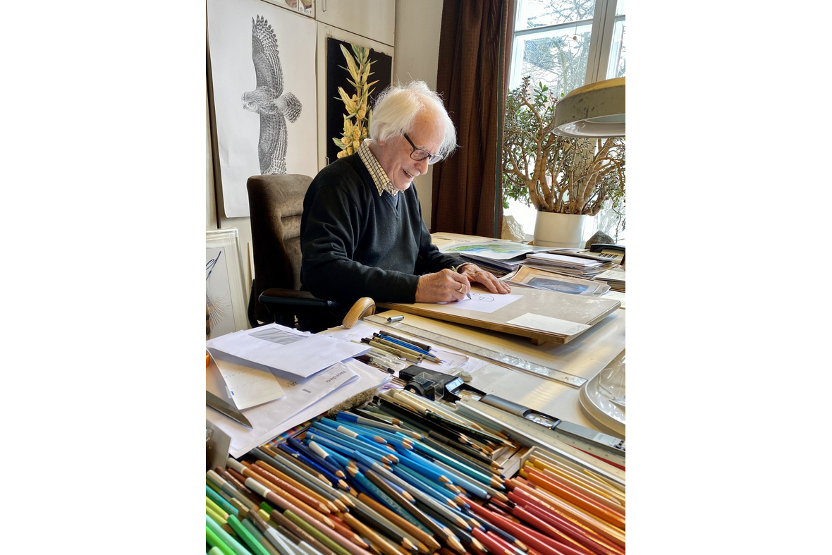 Vanha, harmaahiuksinen mies piirtää pöydän ääressä, jossa paljon eri värisiä kyniä.