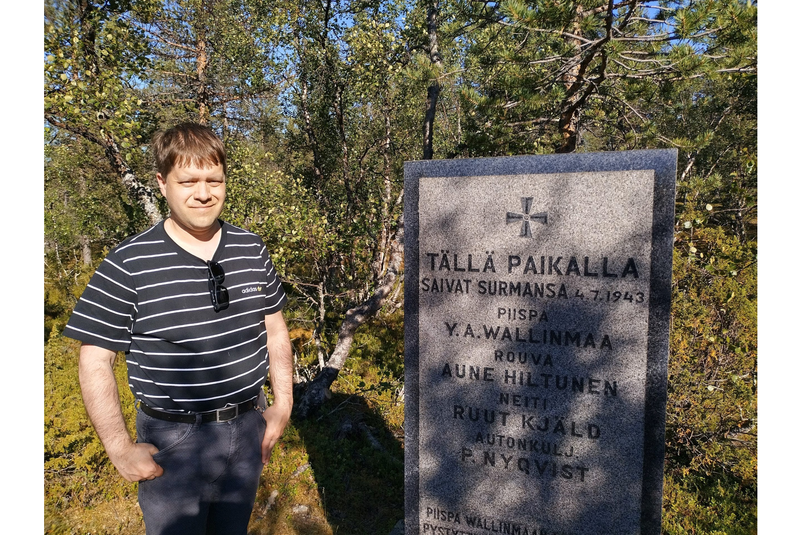 Valokuvassa henkilö seisoo metsäisessä ympäristössä graniittisen muistomerkin vieressä. Muistokiveen on kaiverrettu risti sekä teksti, joka kertoo paikalla surmansa saaneista henkilöistä vuonna 1943. Kiveen on merkitty nimet Y. A. Wallinmaa, Aune Hiltunen, Ruut Kjald ja P. Nyqvist sekä tapahtuman päivämäärä 4.7.1943. Henkilöllä on yllään raidallinen t-paita ja tummat housut. Taustalla näkyy mäntyjä, koivuja ja muuta metsän kasvillisuutta aurinkoisessa säässä. Muistomerkki ja sitä ympäröivä luonto muodostavat kuvan keskeisen aiheen.