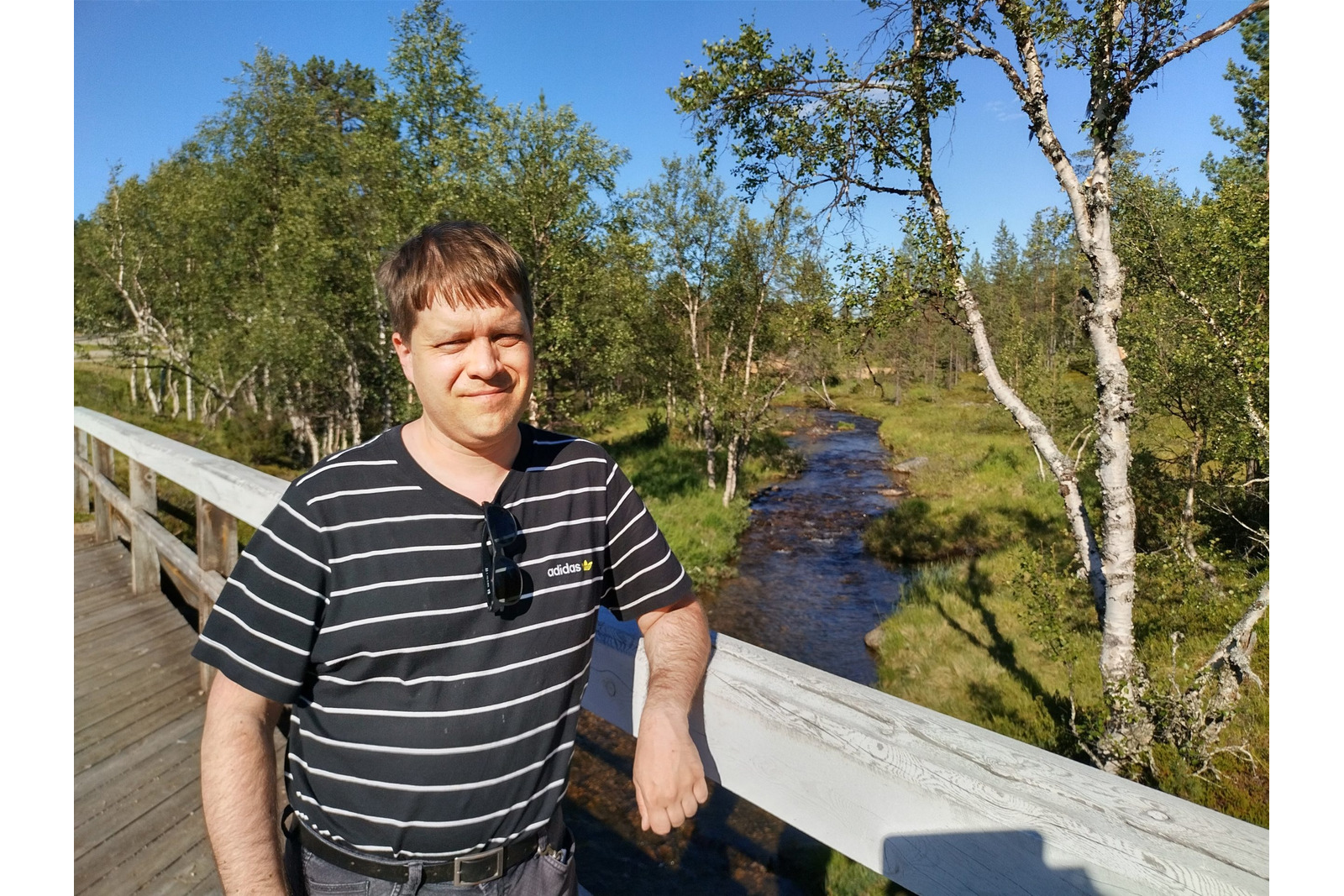 Valokuvassa henkilö nojaa puiseen kaiteeseen sillalla tai pitkospuumaisella kulkuväylällä luonnon keskellä. Taustalla virtaa kapea puro tai joki, jota ympäröivät vihreät koivut, pensaat ja muu kasvillisuus. Henkilöllä on yllään musta-valkoraidallinen t-paita, ja aurinkolasit roikkuvat paidan kaula-aukossa. Päivä on aurinkoinen ja taivas kirkkaan sininen. Kuvan pääosassa ovat sillalta avautuva jokimaisema sekä sitä ympäröivä pohjoiselle luonnolle tyypillinen metsäinen ympäristö.