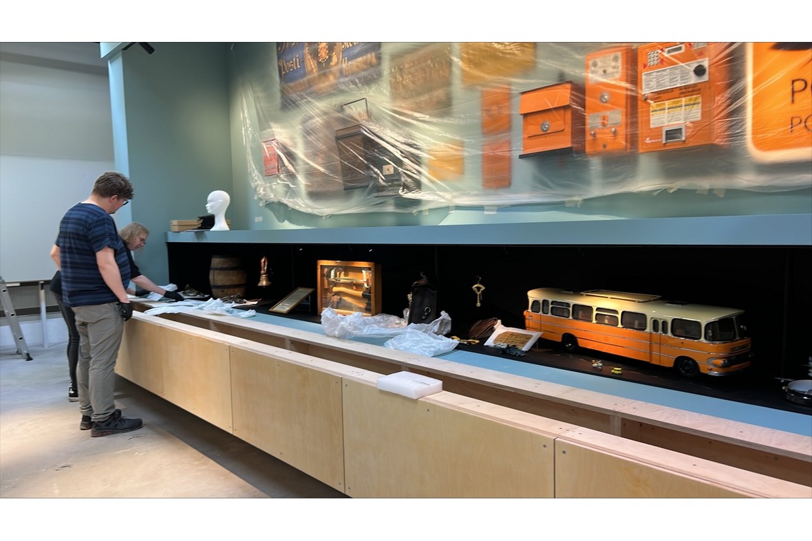 kaksi ihmistä museon näyttelytilassa asettelemassa esineitä, seinällä ja pöydällä jo erilaisia esineitä.