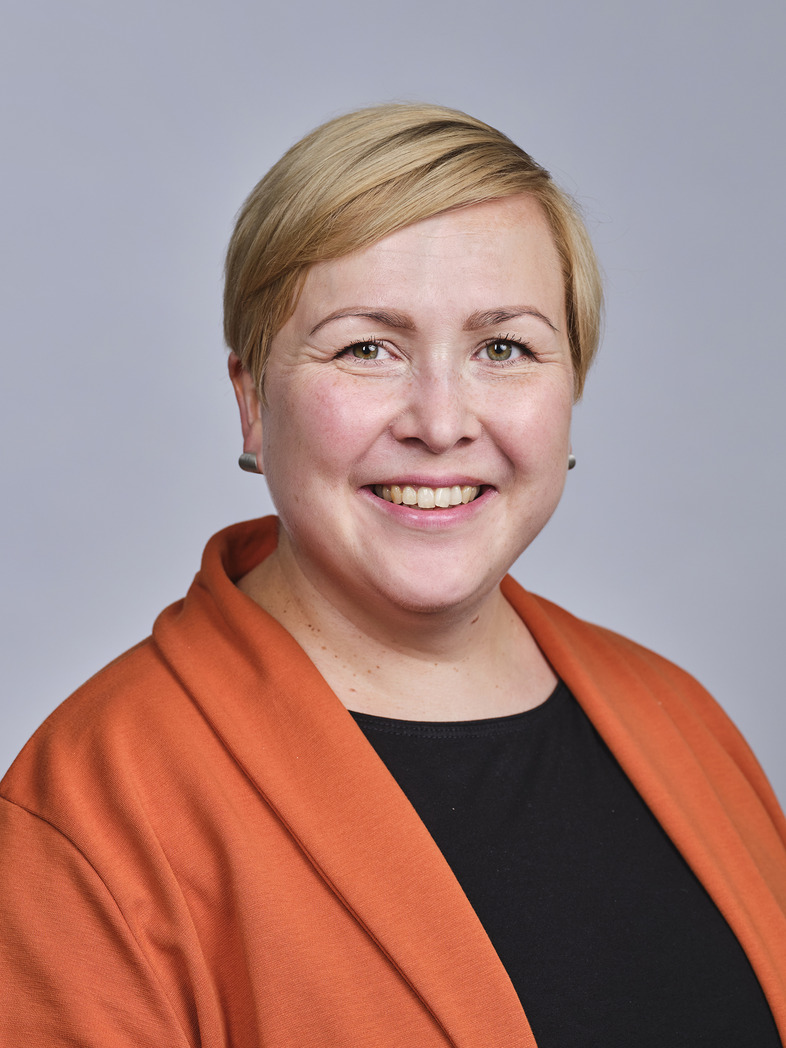 Nainen, lyhyet vaaleat hiukset, oramssi jakki, alla musta paita, hymyilee.
