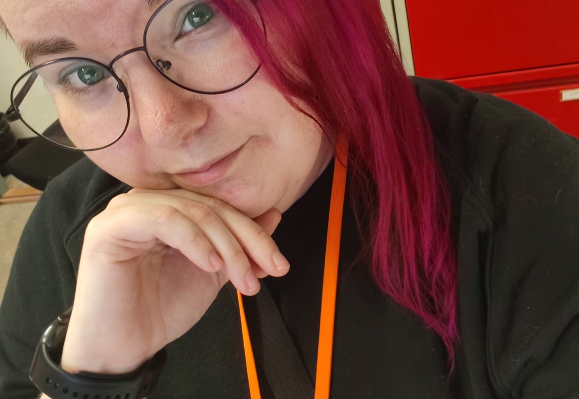 Nuori nainen, pinkki tukka, oranssi avainkaulanauha, musta paita, silmälasit.