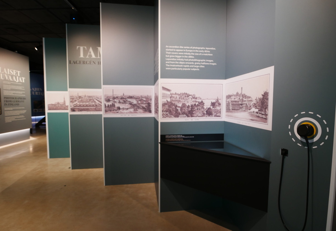 Näyttelytila, jossa oikealla puolella haitarimallinen kuvaseinä, jossa postikorttisuurennoksia ja kuuloke.