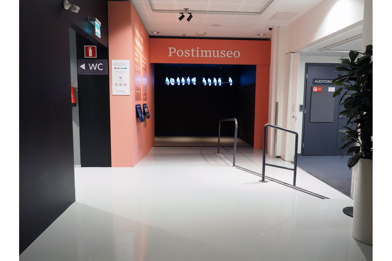 Käytävä, joka päättyy vaaleanpunaiseen sisäänkäyntiin. Ylhäällä teksti Postimuseo. Käytävän päässä mustaa ja kyyhkysen kuvia seinällä. Käytävän puolivälissä on kulut auditorioon ja WC-käytävälle.