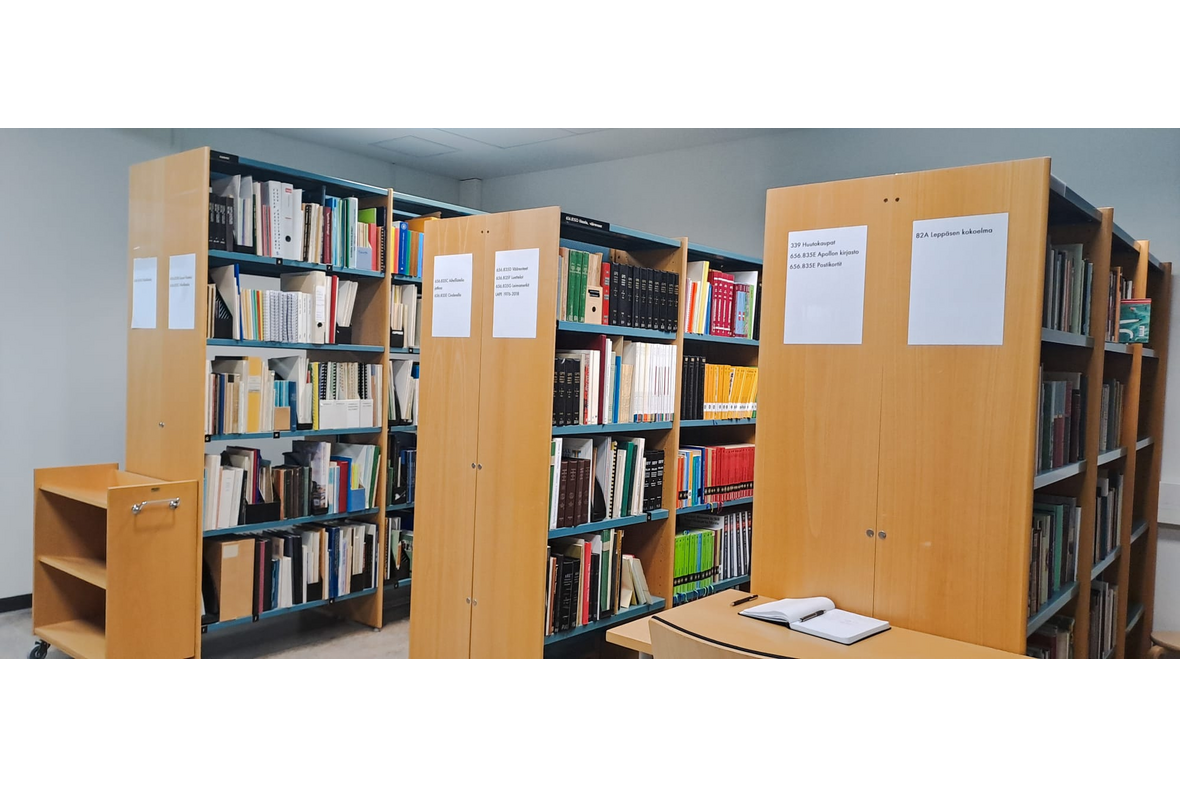 Tila, jossa on hyllykköjä täynnä kirjoja, kirjakärry toisen hyllykköparin sekä pötä ja tuoli toisenb hyllykköparin päädyssä.