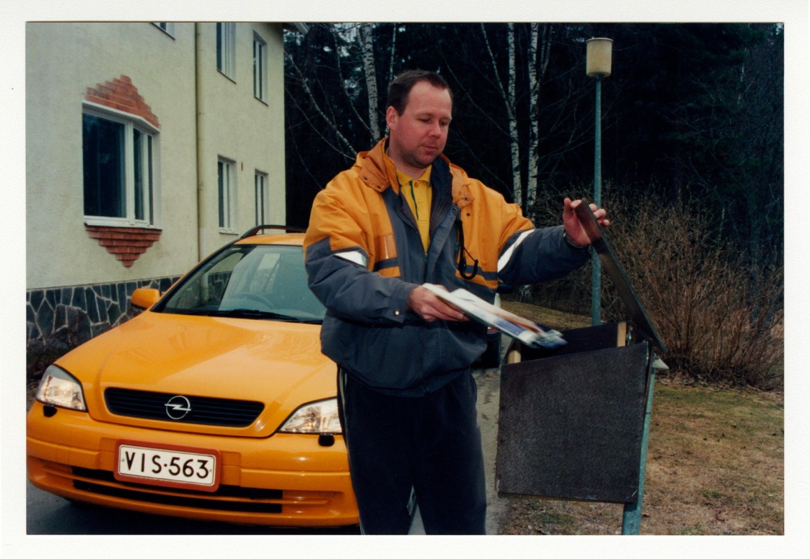 Postinjakaja laittamassa postia postilaatikkoon. Taustalla on oranssin värinen Opel-merkkinen postin jakeluauto.