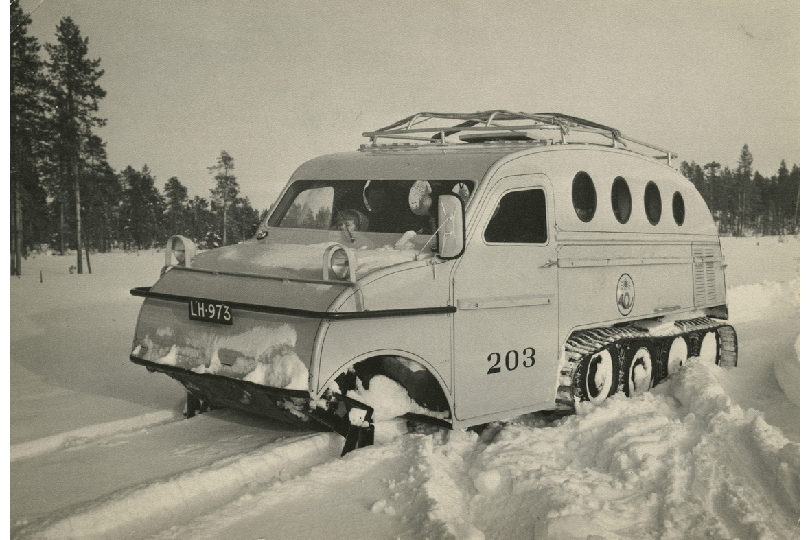 Mustavalkoisessa kuvassa kaareva kattoinen ja telaketjuinen kulkuväline lumisessa metsässä.