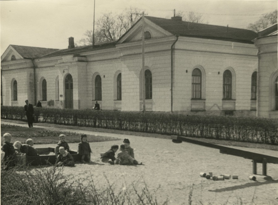 1800-luvun rakennus kaari-ikkunoineen ja pensasaita. Edessä hiekkalaatikolla lapsia leikkimässä.