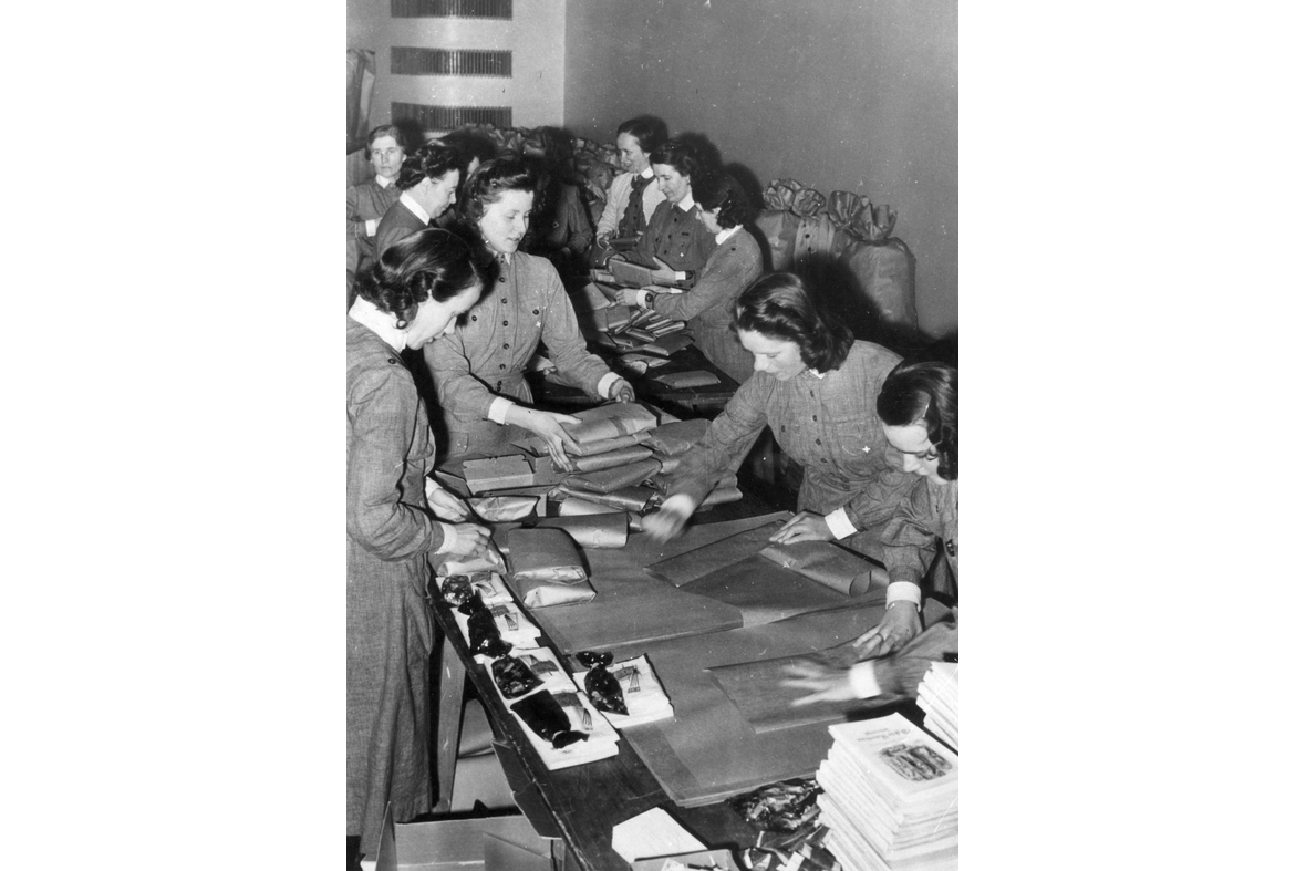 Mustavalkoisessa kuvassa samoihin asuihin pukeutuneet naiset paketoivat tavaroita pitkän pöydän ääressä.