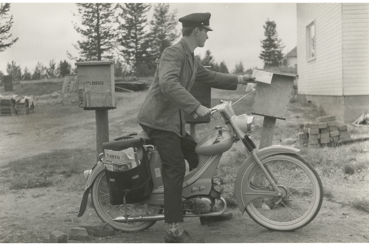 Mustavalkoisessa kuvassa mies virkapuvussa on laittamassa postilaatikkoon kirjettä mopedin päältä. Mopedin takana postilaukku.