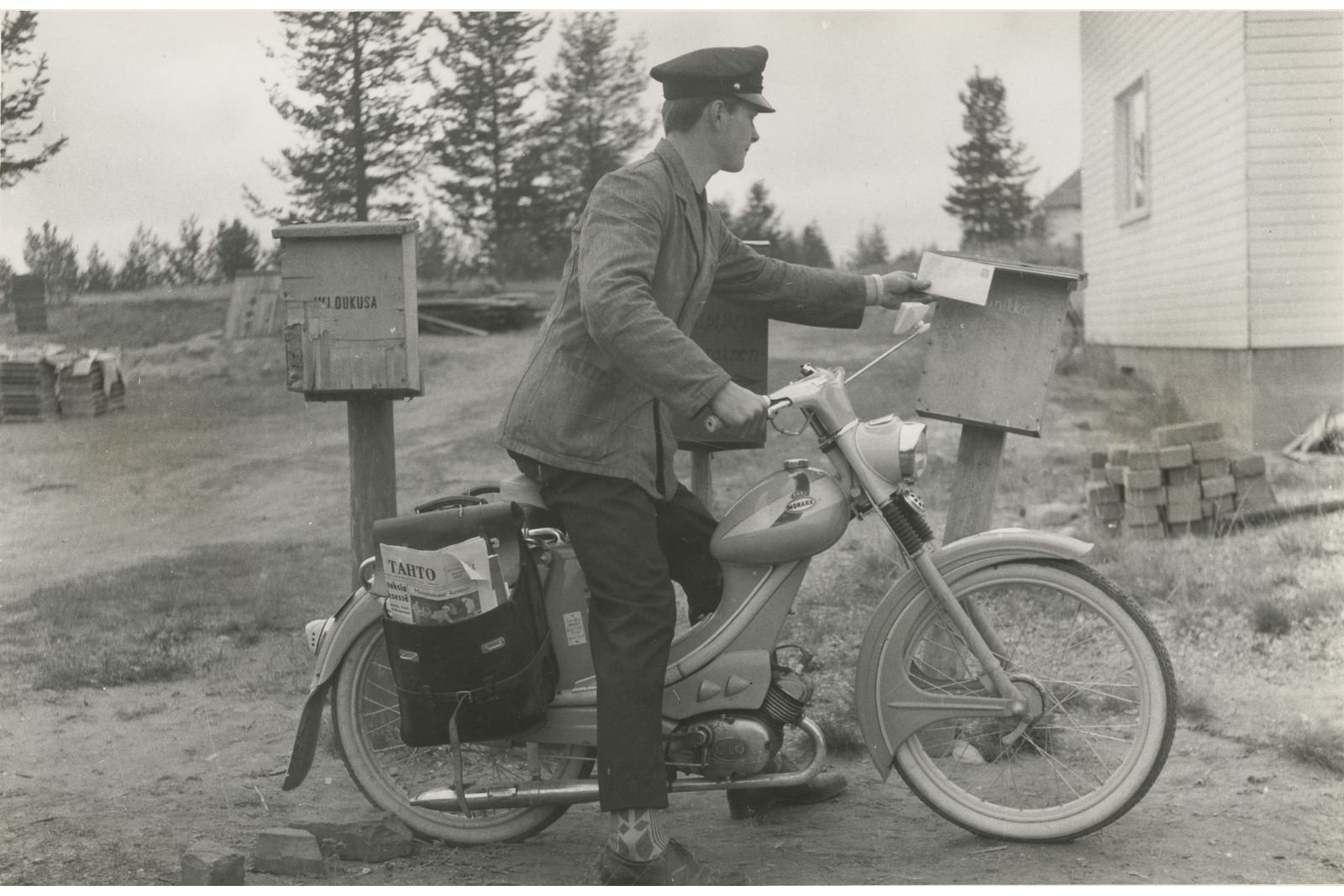 Mustavalkoisessa kuvassa mies virkapuvussa on laittamassa postilaatikkoon kirjettä mopedin päältä. Mopedin takana postilaukku.