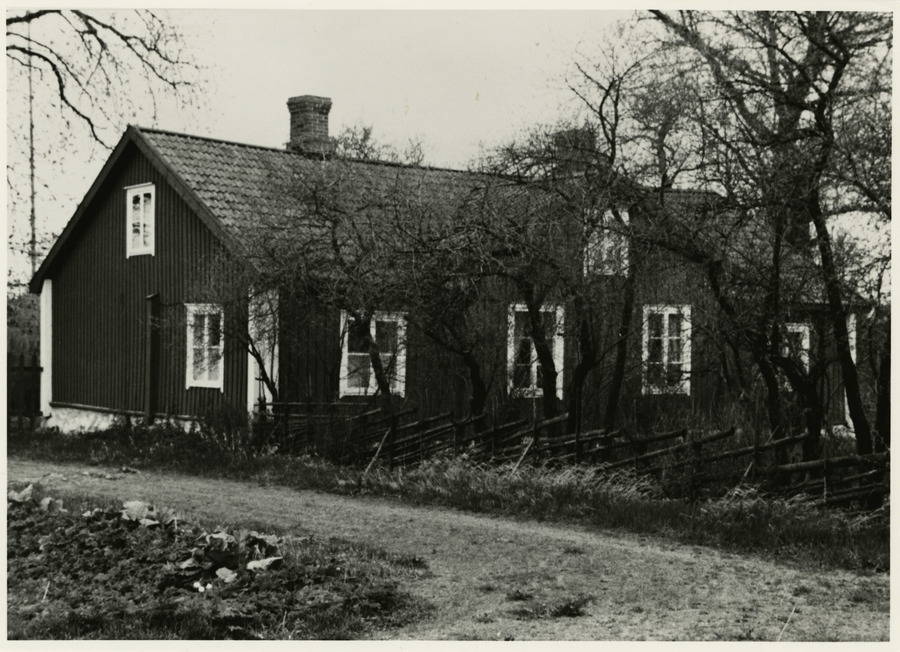 Kapean hiekkatien vieressä on tumma puinen rakennus, jossa on pitkällä sivulla neljä ristikkoikkunaa, päädyssä yksi ja ylhäällä molemmilla sivuilla yksi. Riukuaita alkaa talon kulmalta reunustaen tietä.