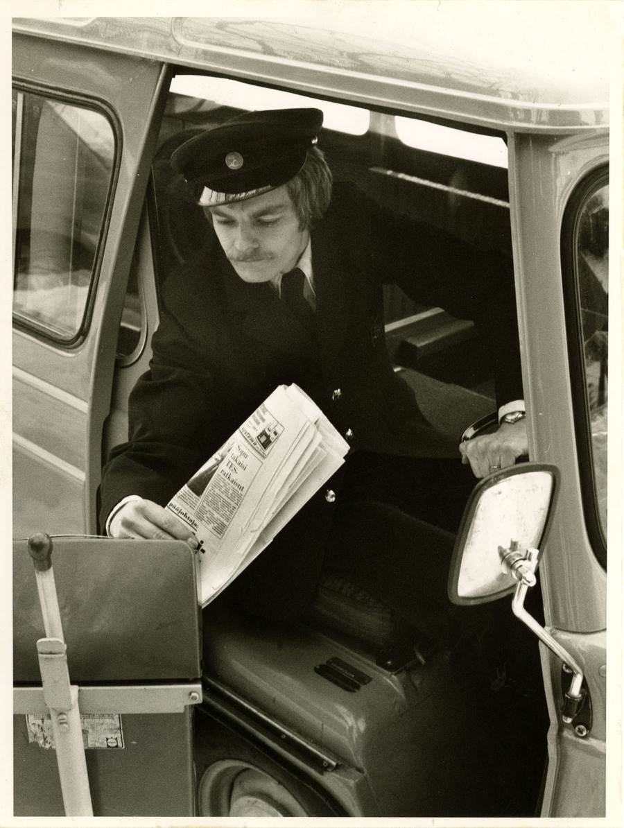 Virkapuvussa oleva postinkuljettaja laittaa sanomalehden postilaatikkoon.