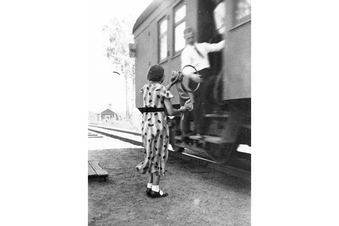 Mustavalkoisessa kuvassa liikkuvan junan rappusilta mies on tarttumassa vanteeseen, jossa on kiinni postipussi. Pussia asemalla pitelee nainen mekossa.