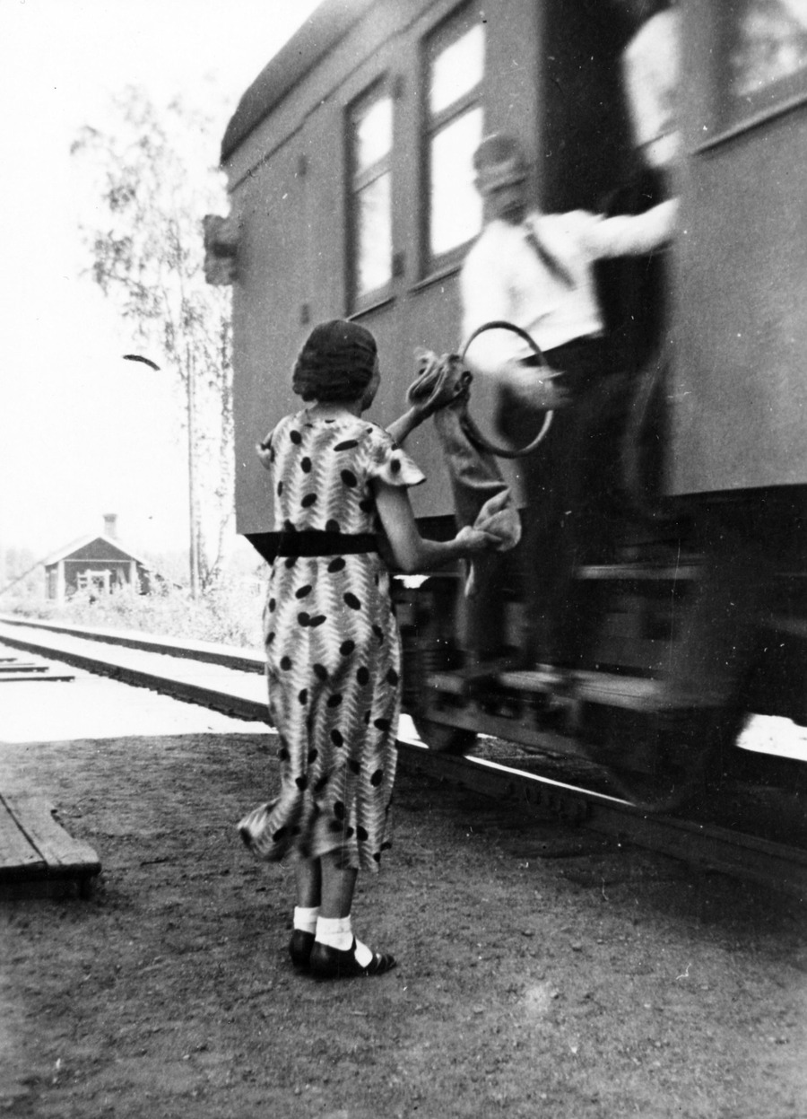 Mies ojentaa liikkuvan junan oviaukosta postipussin ulkona seisovalle naiselle.