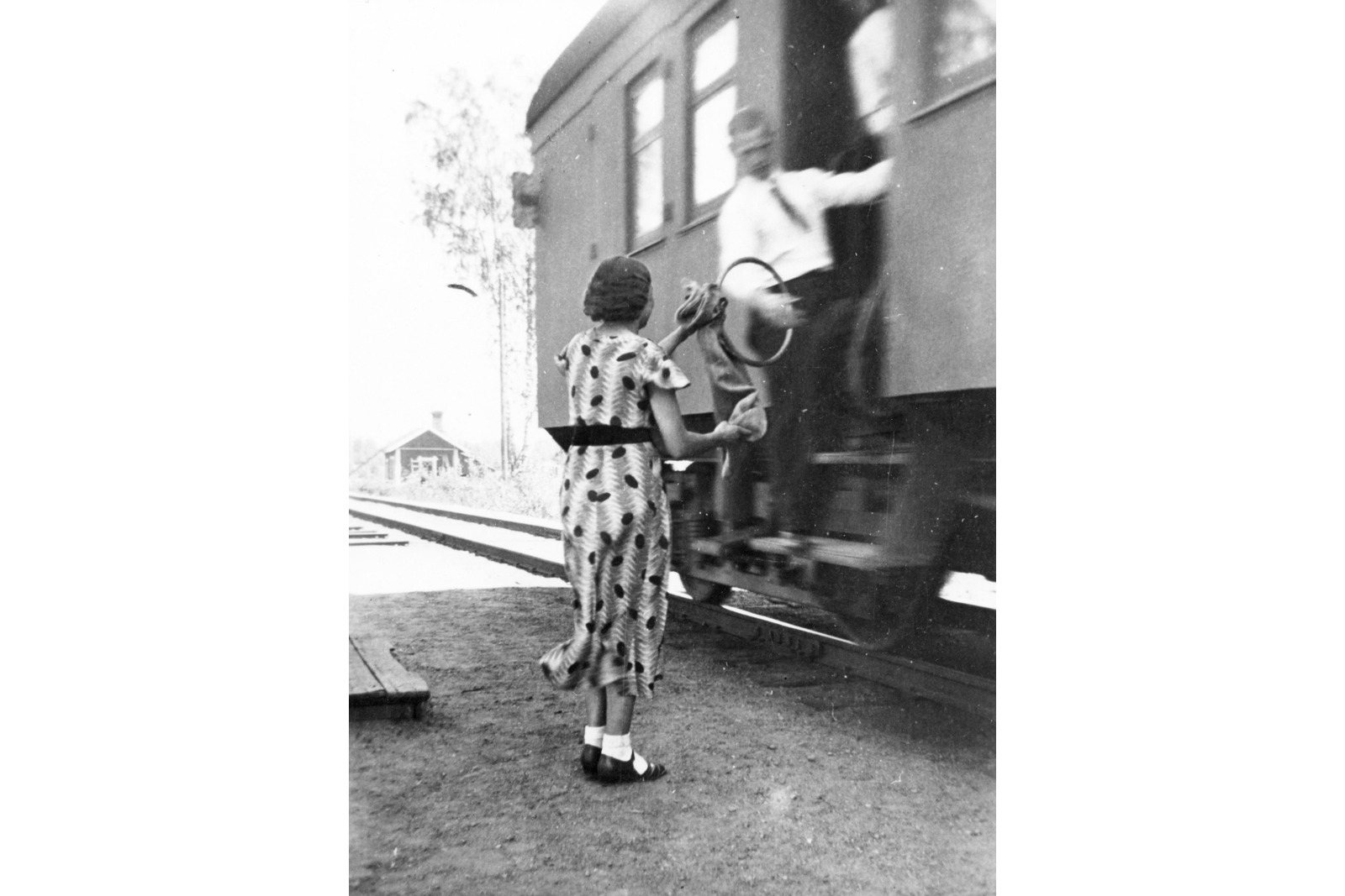 Mustavalkoisessa kuvassa liikkuvan junan rappusilta mies on tarttumassa vanteeseen, jossa on kiinni postipussi. Pussia asemalla pitelee nainen mekossa.