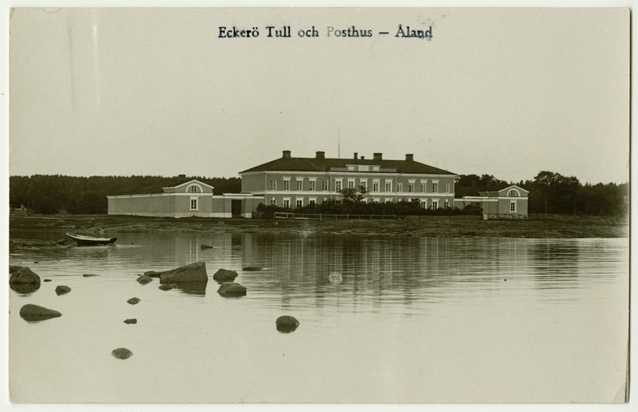 Mustavalkoisessa kuvasssa vesialueen vastarannalla empiretyylinen rakennus. Kuvan yläpuolella teksti Eckerö Tull och Posthus - Åland.