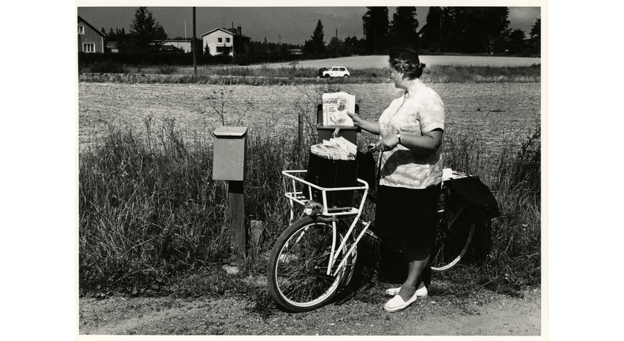 Nainen napitettavassa lyhythihaisessa paidassa ja hameessa polkupyörällä laittamassa sanomalehteä postilaatikkoon, taustalla peltoa, maantie ja pari taloa.