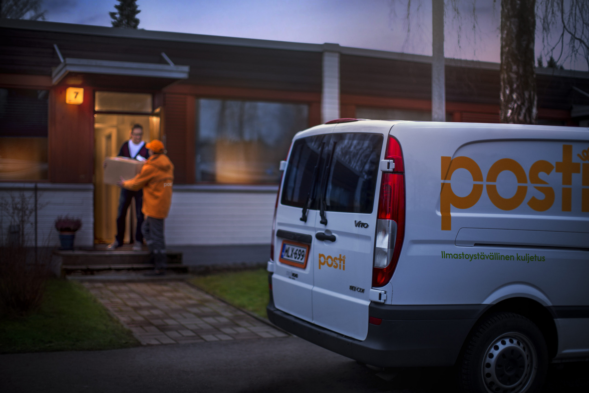Postinjakaja ojentamassa ovella pakettia asiakkaalle, tien vieressä Postin pakettiauto.
