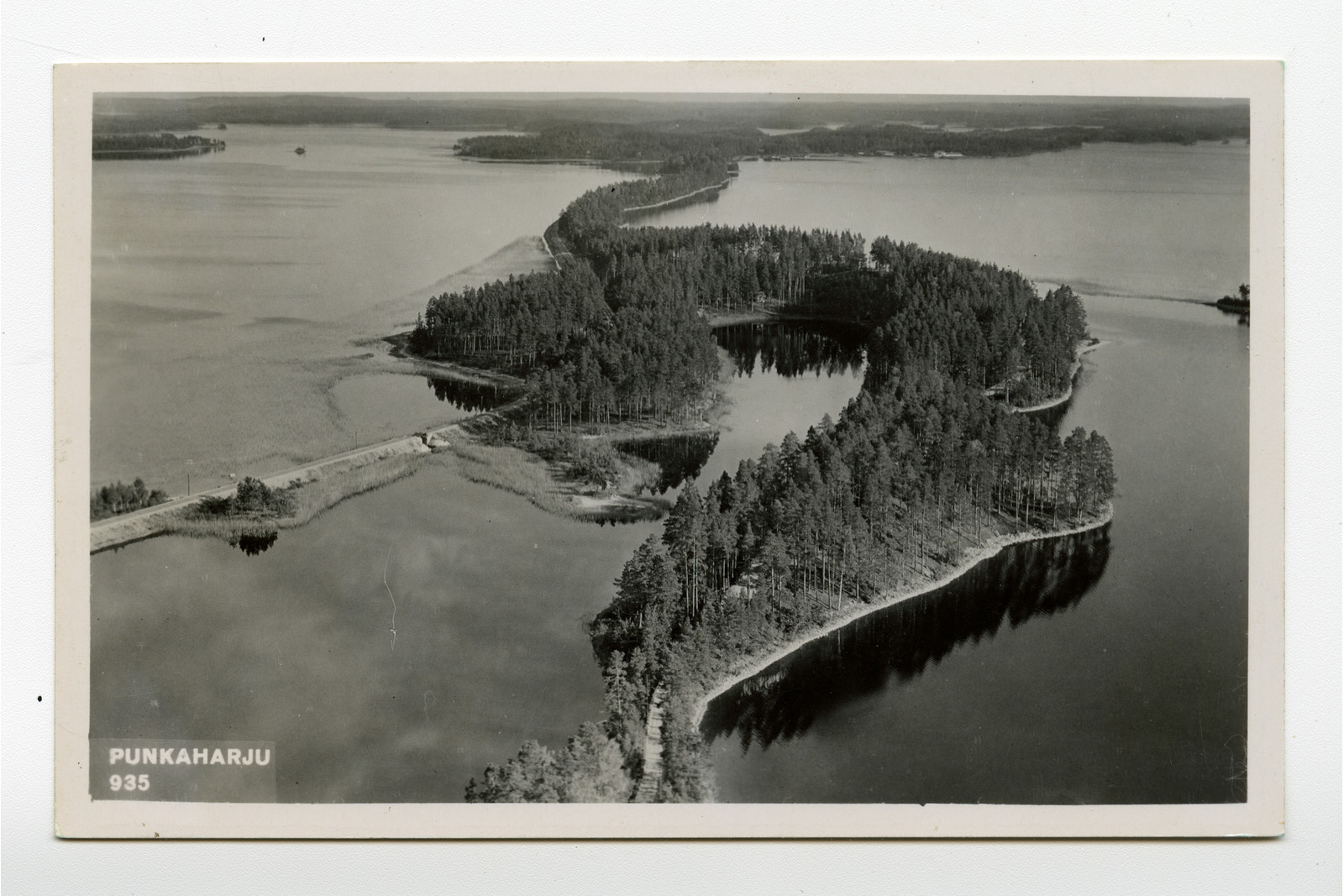 Mustavalkoinen vaakatasoinen postikortti, jossa on kuva järvestä, jota halkoo kiemurteleva maavana teineen. Vasemmalla alakulmassa teksti Punkaharju 935.