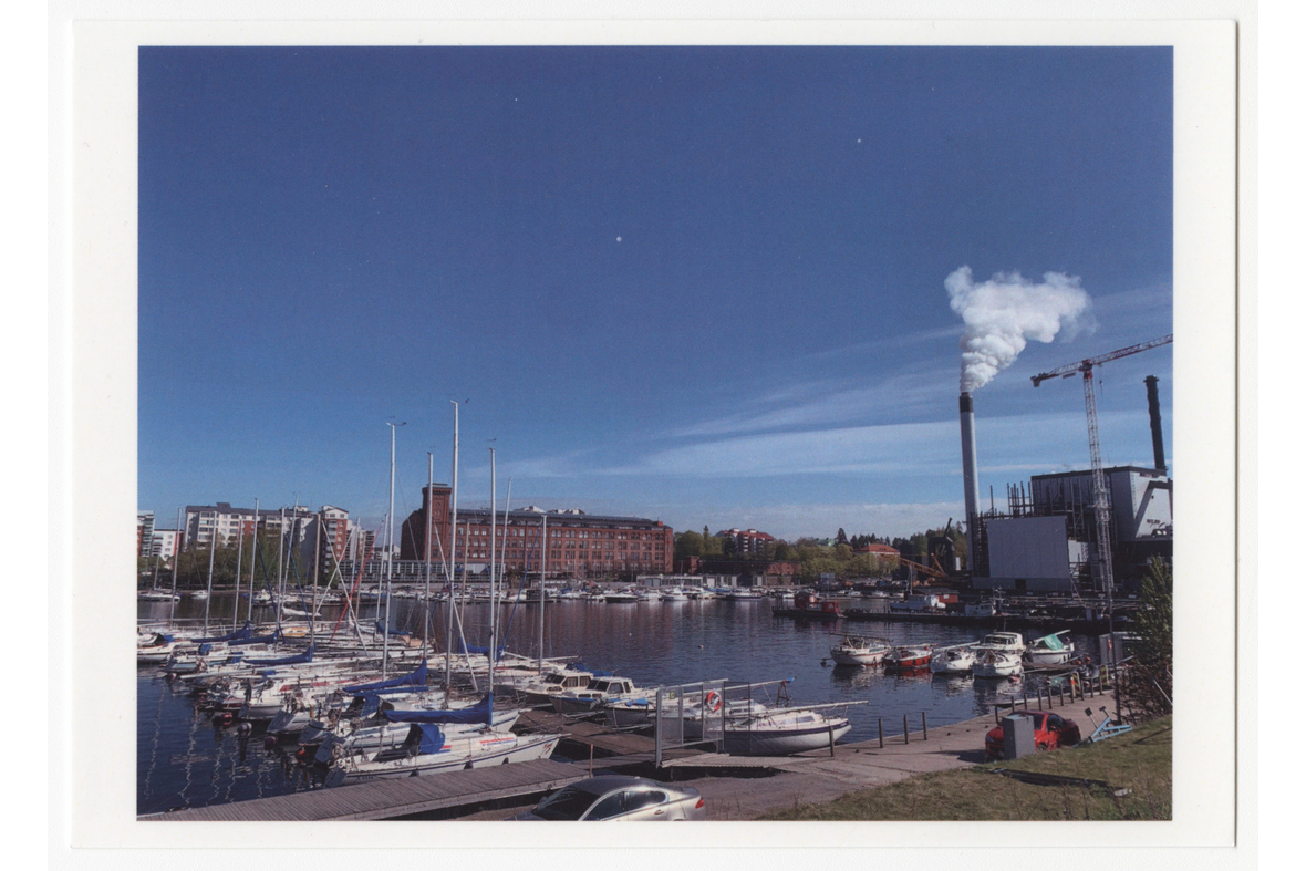 Vaakatasoinen postikorttikuva järven rannasta venelaitureineen, vasemmalla voimalaitos ja vastarannalla vanha tiilinen tehdasrakennus sekä kerrostaloja.