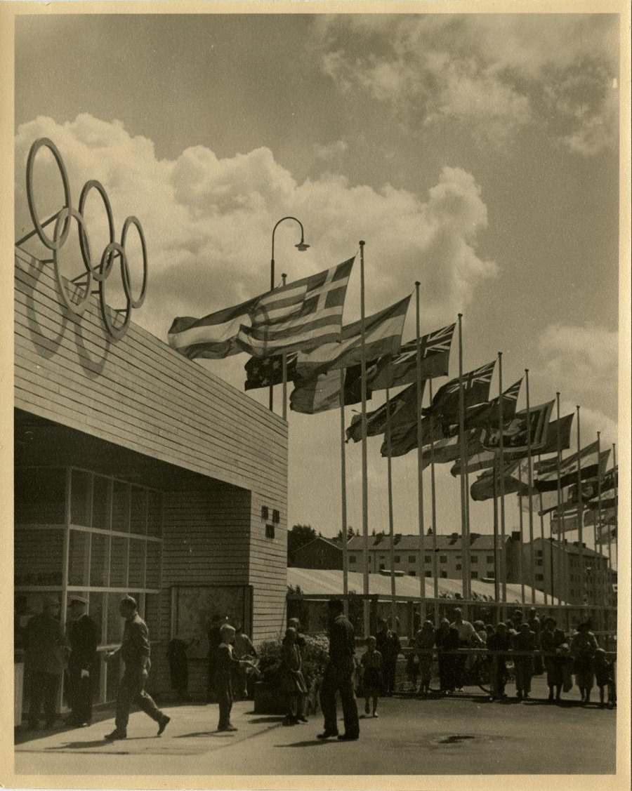sisällön kuvaus: Helsingin olympialaiset. Kisaportti Käpylän kisakylässä. Eri maiden lippuja, urheilijoita ja lapsia portin edessä ja aidan takana ihmisiä. Helsingin olympialaiset järjestettiin 19.7.-3.8.1952.