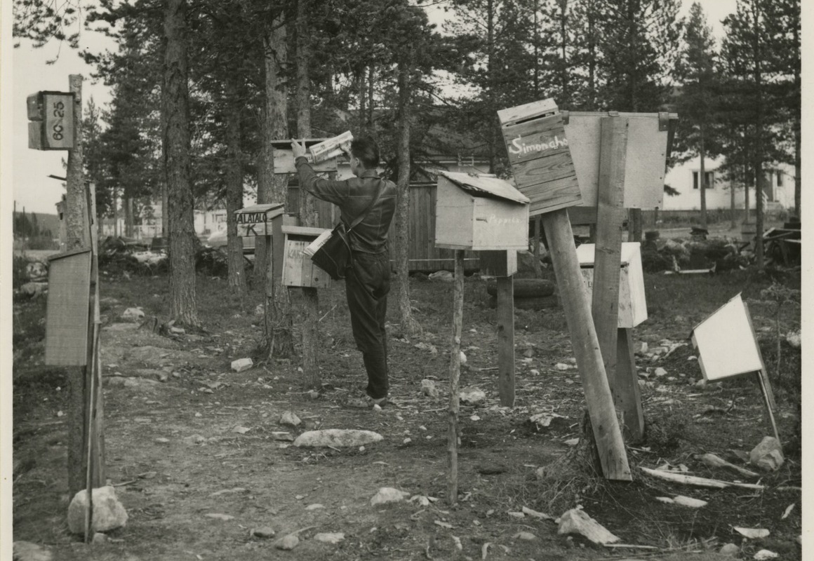 Mustavalkoisessa kuvassa jakelija on laittamassa sanomalehteä puussa olevaan postilaatikkoon, ympärillä hajanaisesti erilaisia postilaatikoita useiden eri tolppien päässä metsän reunassa, talo puiden takana.