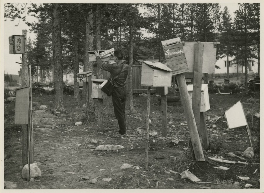 Mustavalkoisessa kuvassa jakelija on laittamassa sanomalehteä puussa olevaan postilaatikkoon, ympärillä hajanaisesti erilaisia postilaatikoita useiden eri tolppien päässä metsän reunassa, talo puiden takana.