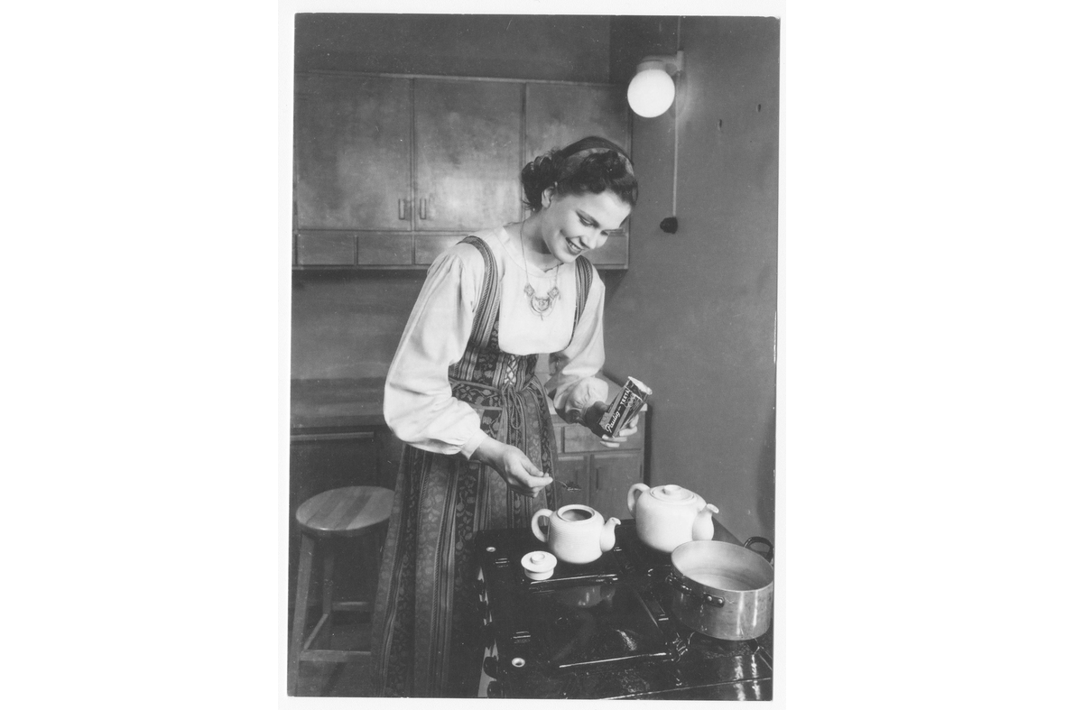 Mustavalkoisessa kuvassa kansallispuvussa nainen keittämässä kahvia puuhellalla.