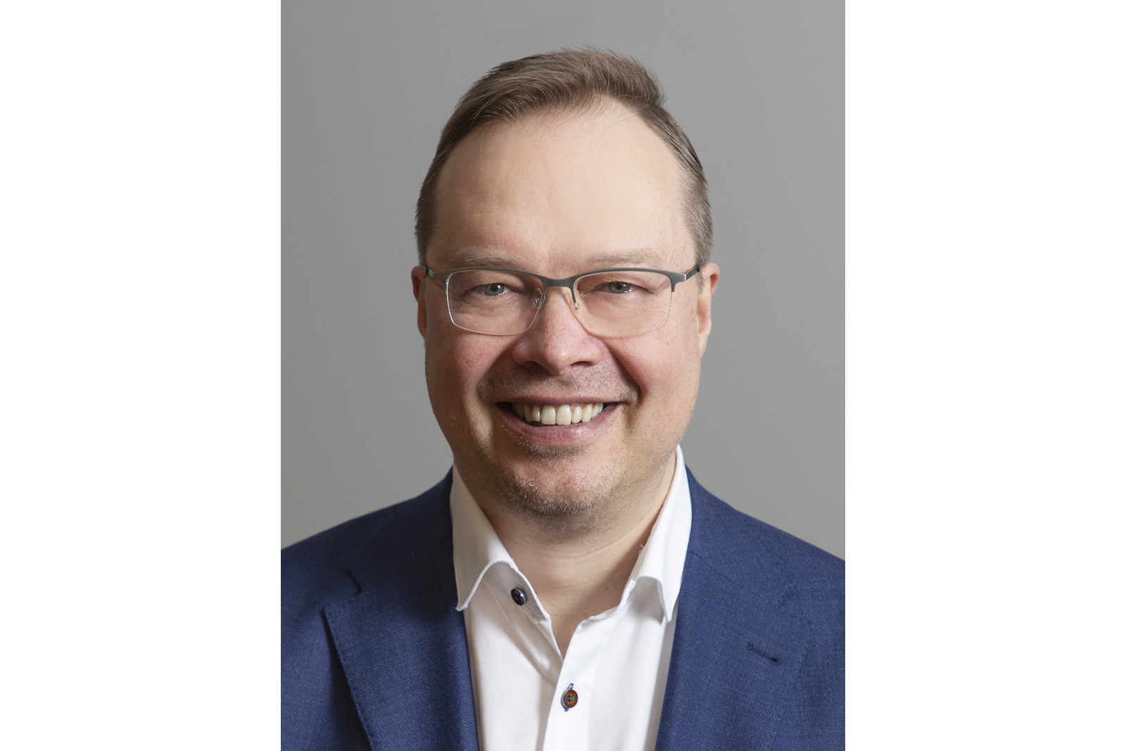 Hymyilevä vaaleahiuksinen ja silmälasipäinen mies kauluspaidassa ja puvun takissa.
