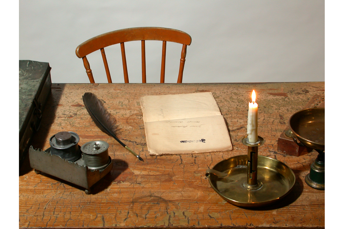 Pöydällä kynttilä palamassa, sulkakynä, paperia ja muita tavaroita, pöydän takana tuoli.