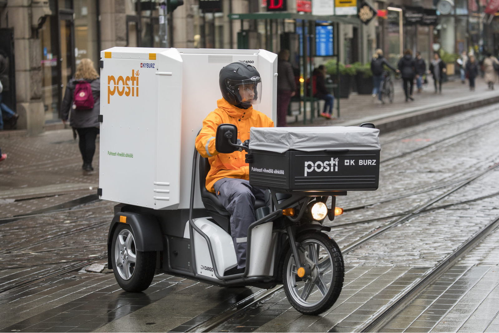 Postinjakaja ajamassa sähkörahtiskootteria raitiotien yli suojatien kohdalla märällä kelillä.