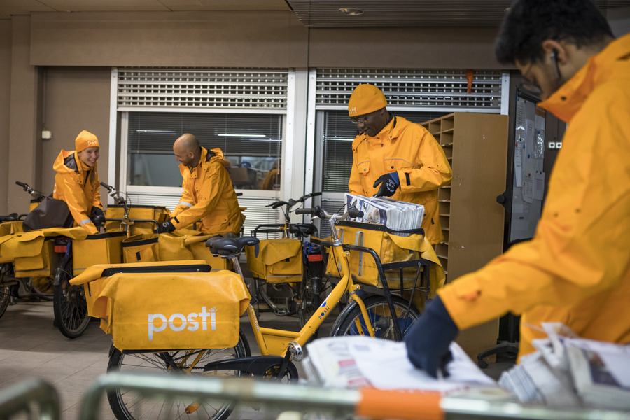 Neljä henkilöä häärii keltaisten Postin logoilla varustettujen polkupyörien ympärillä. Takaritilöillä pyörälaukuissa postia, samoin etukopassa. Niin asut kuin pyörät ovat kokonaisuudessaan keltaisia. Taaimmainen jakaja hymyilee leveästi.