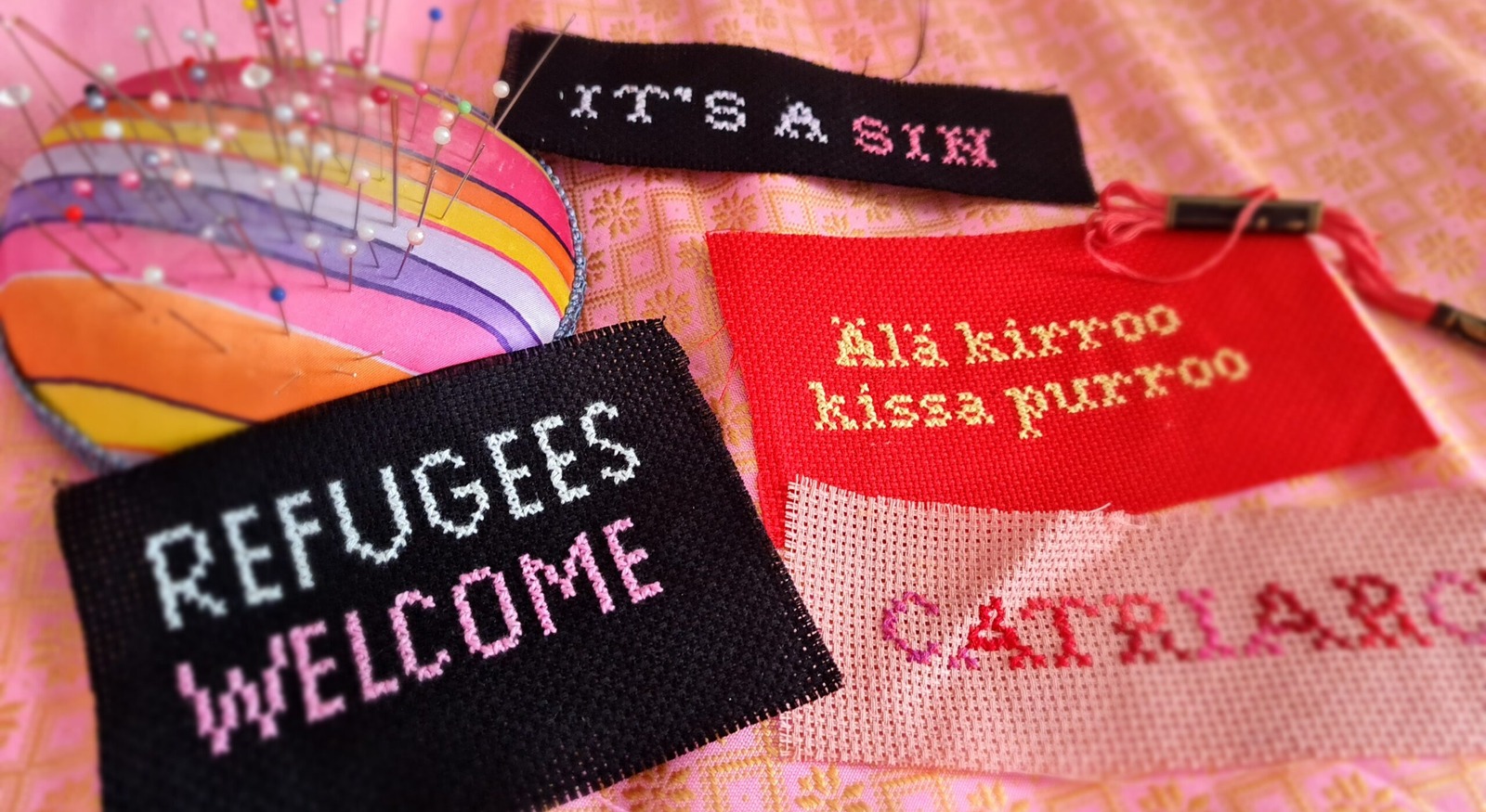 Ristipistoja värikkäillä kankailla. Teksteinä: It´s a sin, Refugees welcome, Älä kirroo, kissa purroo, Catriarc.