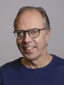 Hymyilevä silmälasipäinen harmahtava hiuksinen mies sinisessä neulepuserossa.