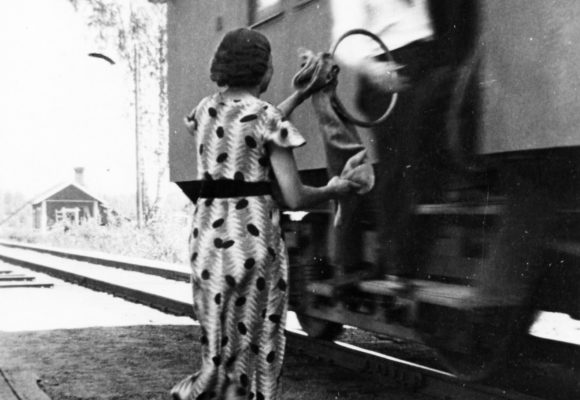 Mies ojentaa liikkuvan junan oviaukosta postipussin ulkona seisovalle naiselle.