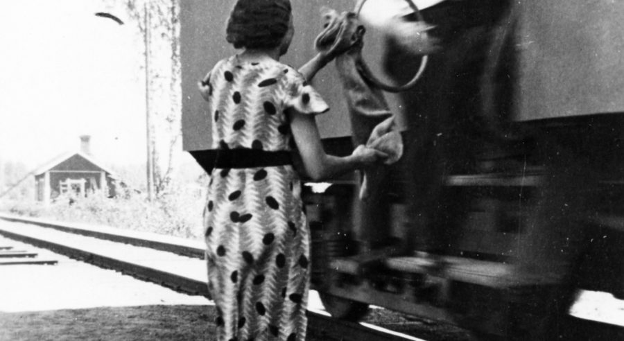 Mies ojentaa liikkuvan junan oviaukosta postipussin ulkona seisovalle naiselle.