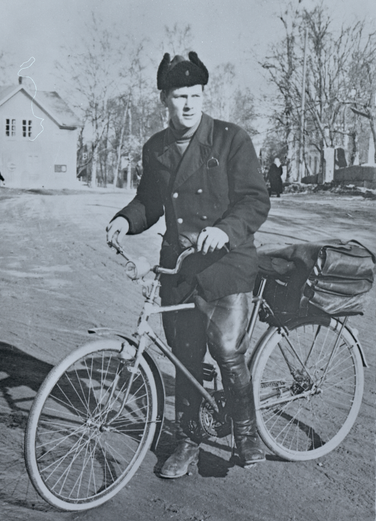 Mustavalkoisessa kuvassa postinjakaja pyörän selässä, päällään turkislakki, päällystakki, nahkahousut ja -saappaat.