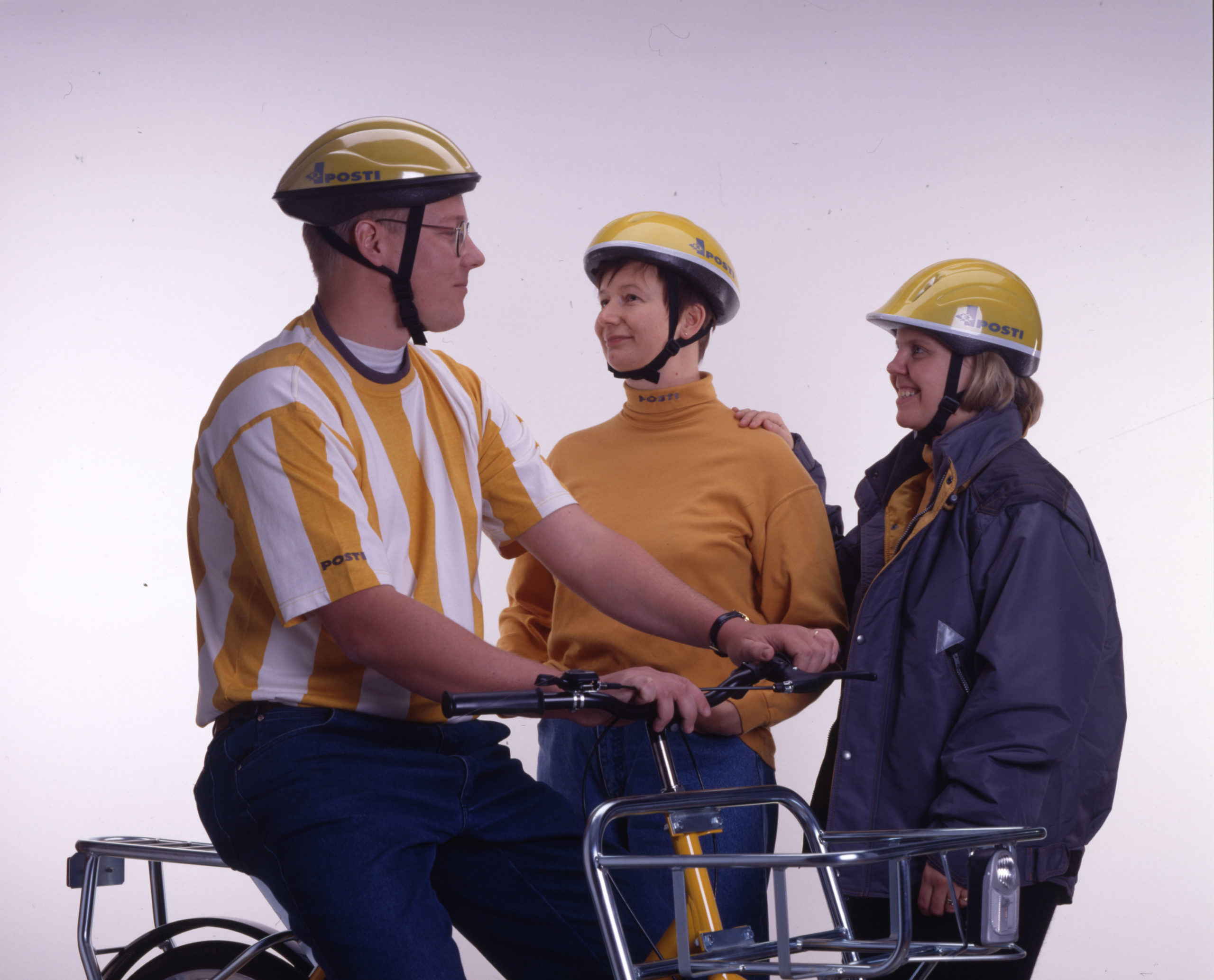 Kolme ihmistä postinjakajien vuoden 1996 asuissa. Jokaisella on keltainen kypärä. Jakelupyörän selässä olevalla on kelta-valkopystyraidallinen t-paita, vieressään olevalla keltainen kauluspusero ja reunimmaisilla harmaa kansitakki.